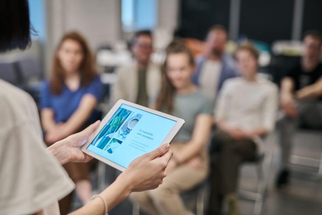 Eine Person hält ein Tablet mit einer Präsentation, während eine Gruppe von Menschen im Hintergrund in einem Seminarraum oder Meeting aufmerksam zuhört.
